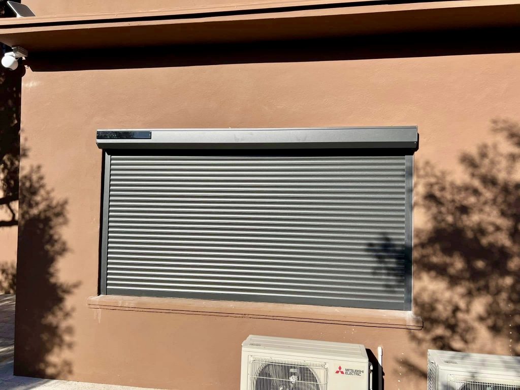 Volet roulant aluminium gris sur façade ocre avec climatiseur extérieur
